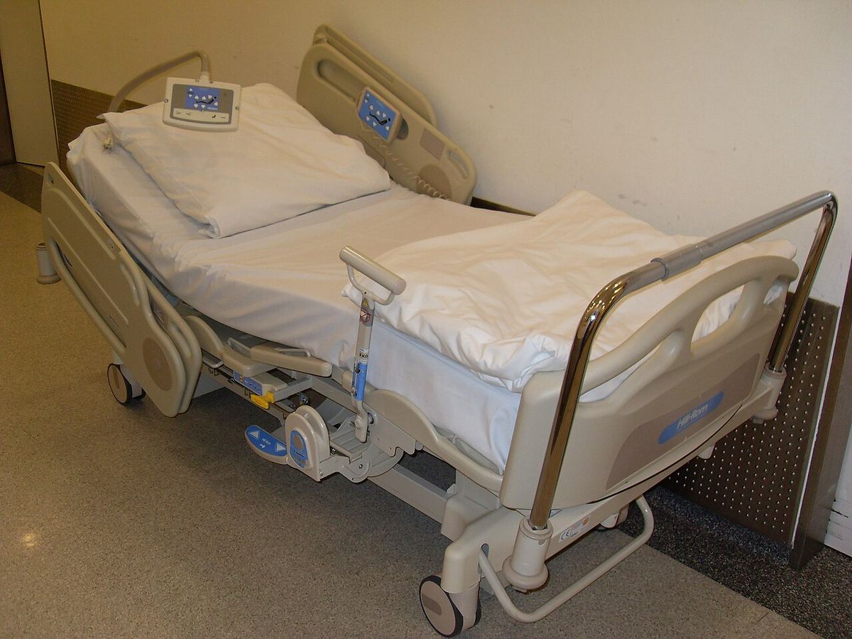 An empty electric hospital bed with white sheets, side rails and a handheld control, parked against a wall