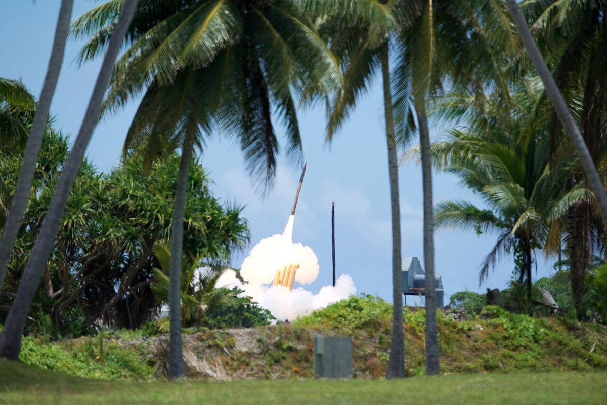 A THAAD interceptor missile launches in a plume of white smoke behind tall palm trees under a blue sky