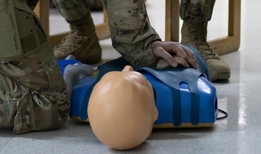 Hands press down on the chest of a blue CPR training manikin on a classroom floor