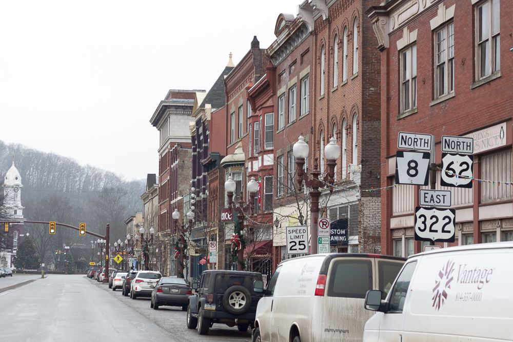 Brick storefronts and parked cars along a main street in downtown Franklin, Pennsylvania, with wooded hills behind