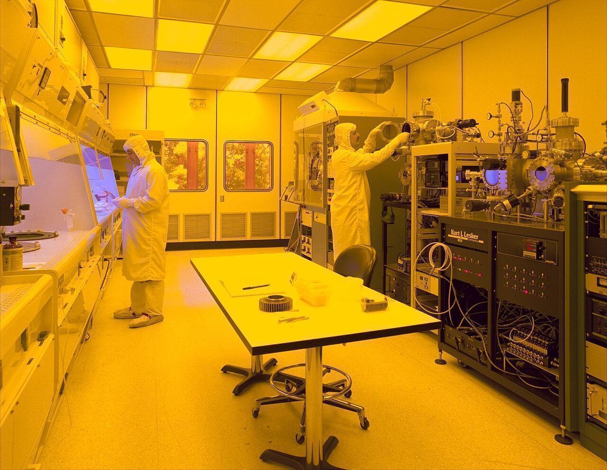 Workers in full-body cleanroom suits at equipment benches in a yellow-lit microfabrication cleanroom