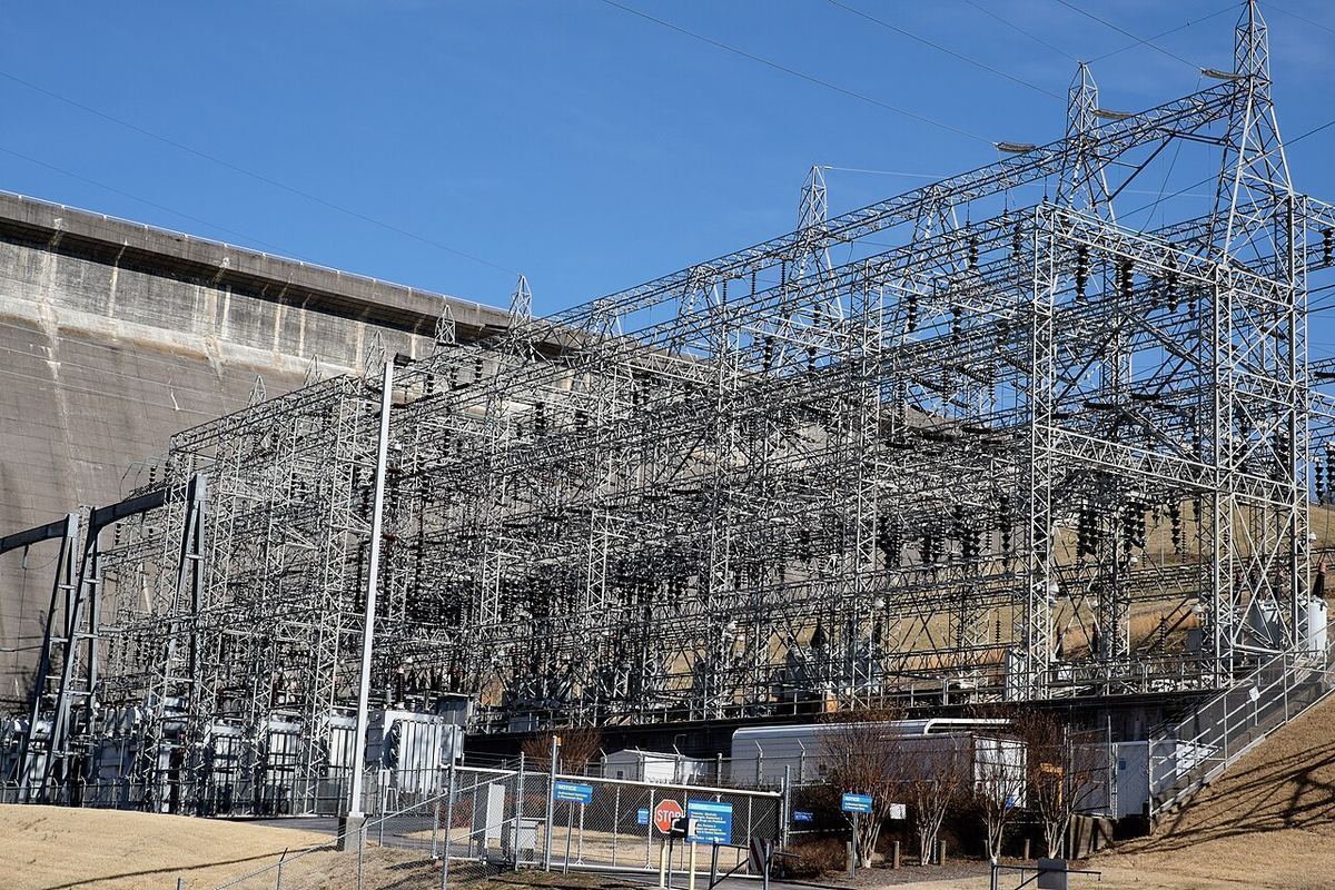 Steel towers and equipment of an electrical substation beside a concrete dam under a clear blue sky