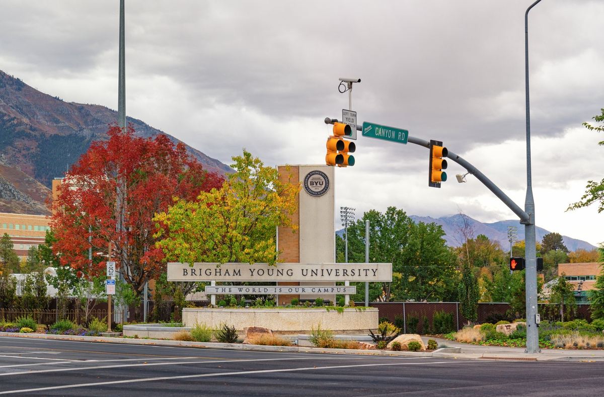 The Brigham Young University entrance sign framed by red and gold autumn trees, with mountains behind